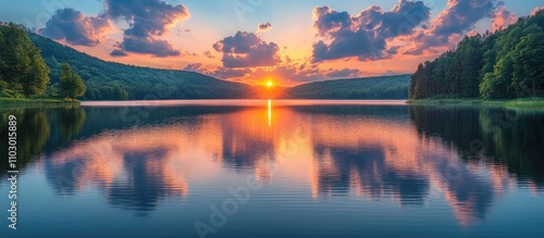 Fototapeta Naklejka Na Ścianę i Meble -  Peaceful sunset over serene lake with colorful cloud reflections calm water lush green hills and trees in the background during early summer