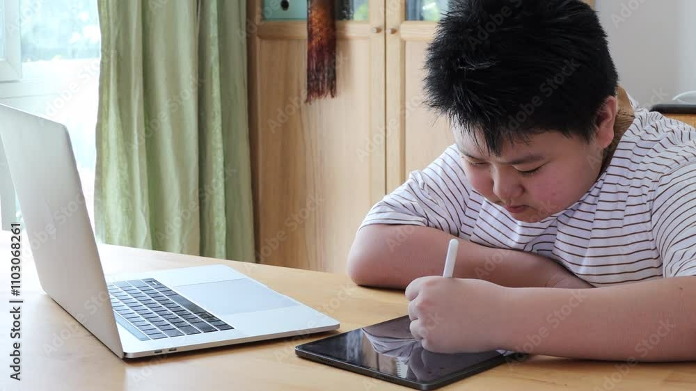 A young Asian boy using a digital tablet and laptop to do homework at home during the daytime. A young Asian boy using a digital tablet and laptop to do homework at home during the daytime.