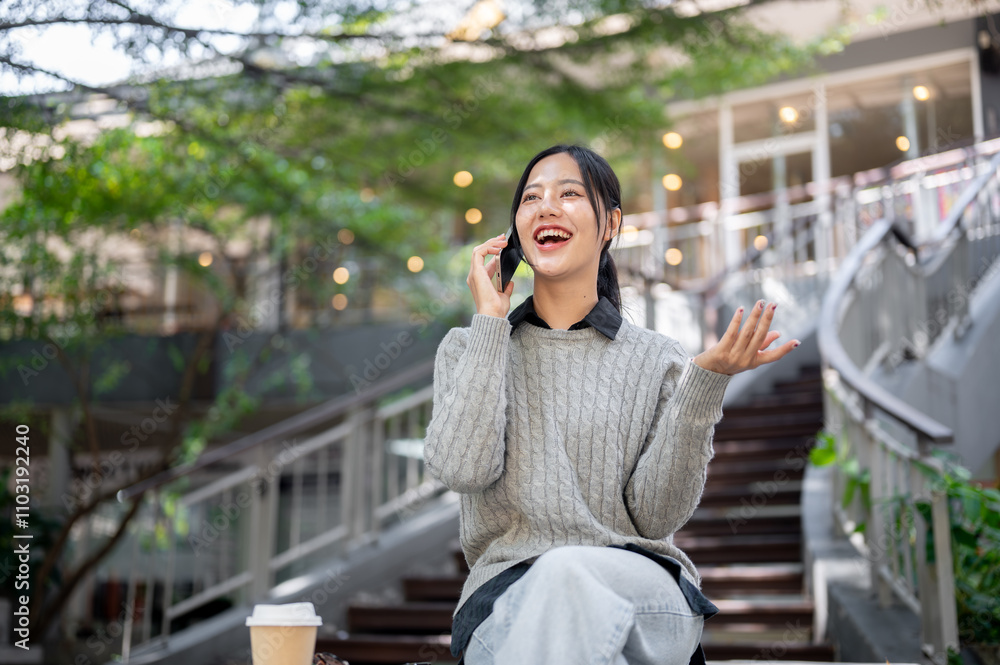Fototapeta premium A cheerful, smiling Asian woman enjoys talking on the phone while sitting outdoors in the city.