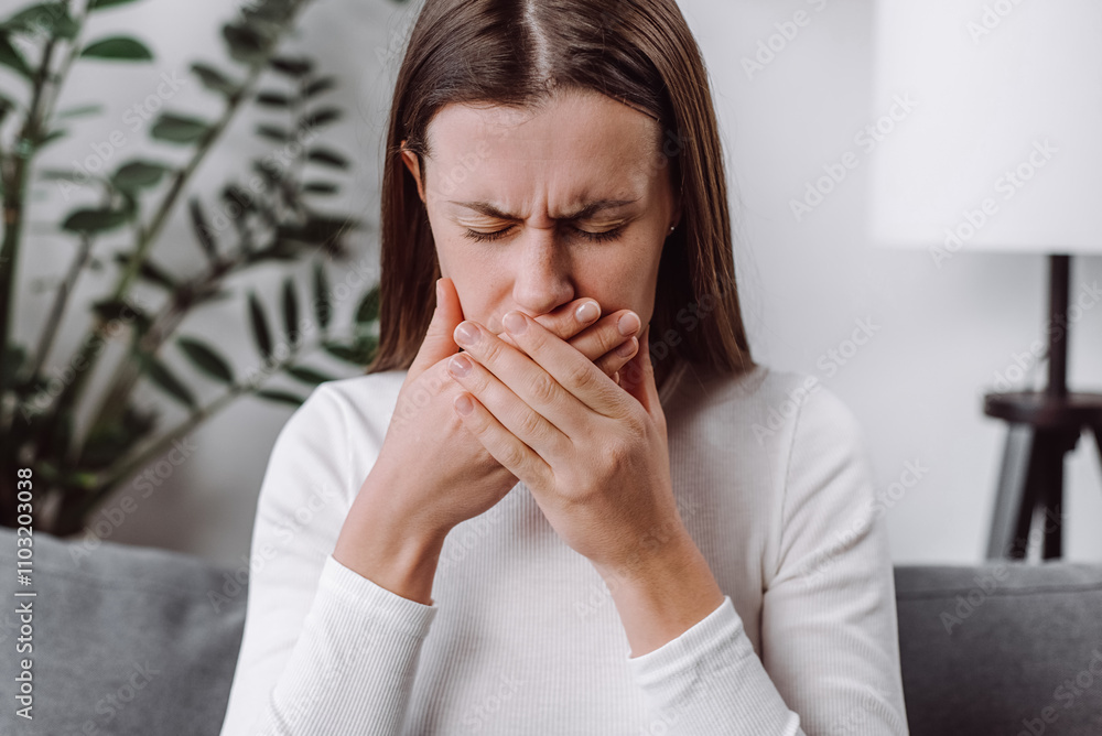 Close up of young woman suffering from nausea at home, caucasian female having morning sickness, ill lady sitting alone on couch in living room, covering mouth Close up of young woman suffering from nausea at home, caucasian female having morning sickness, ill lady sitting alone on couch in living room, covering mouth