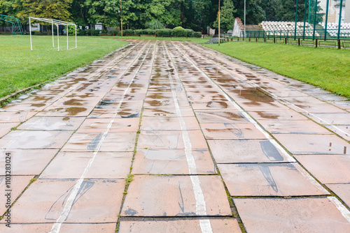 Old damaged running track in stadium with faded white lines and broken tiles, surrounded by green grass and fields. Urban sports facilities concept photo