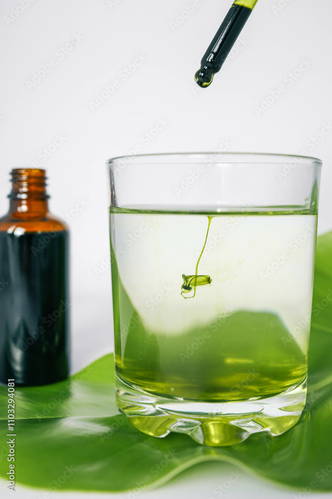 Dropper releasing green liquid into a glass of water with a leaf underneath and a brown glass bottle in the background. Represents natural ingredients, organic products. Dropper releasing green liquid into a glass of water with a leaf underneath and a brown glass bottle in the background. Represents natural ingredients, organic products.