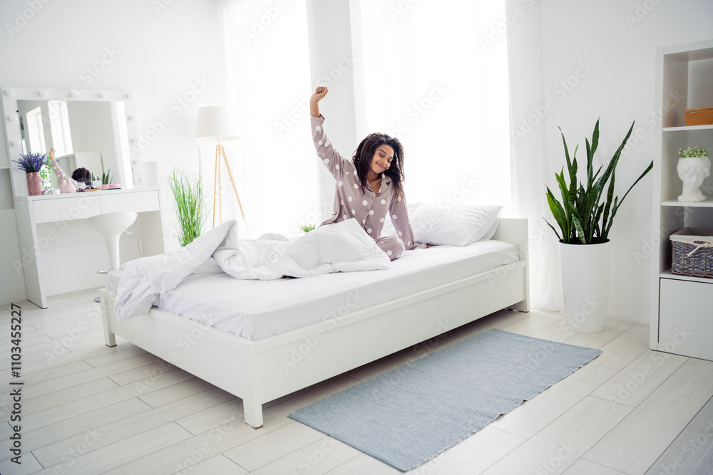 Obraz premium Cheerful afro woman in pajamas waking up in a bright bedroom surrounded by natural light and cozy decor