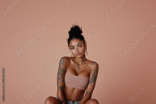 Close-up of a sexy and hot black afro-american woman with a tan complexion and floral tattoos wearing a sleeveless top in skin color, set against a soft pink peachy background photo