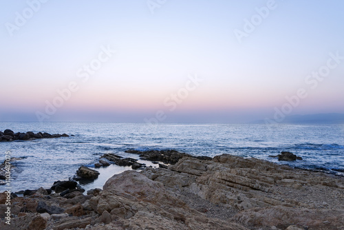 A serene coastal scene at twilight. The rocky shoreline extends into calm waters, reflecting the soft hues of the evening sky. The gradient transitions from light blue to soft pink and purple