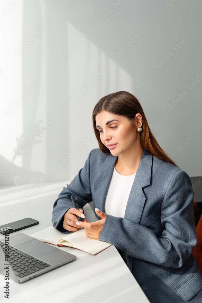A young woman in a gray blazer works on a laptop at a white desk in a minimalist home office. The setup includes a beige notebook, a decorative vase with dried plants, and natural light. A young woman in a gray blazer works on a laptop at a white desk in a minimalist home office. The setup includes a beige notebook, a decorative vase with dried plants, and natural light.