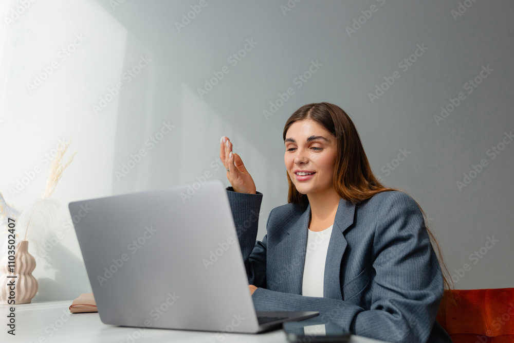 Woman in a gray blazer is looking at her laptop and smiling during an online conference. The workplace is well lit and a vase of dried plants adds aesthetic appeal. Copy space. Woman in a gray blazer is looking at her laptop and smiling during an online conference. The workplace is well lit and a vase of dried plants adds aesthetic appeal. Copy space.