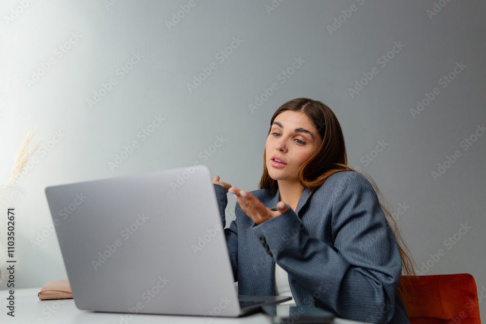 Woman in a gray blazer is looking at her laptop and smiling during an online conference. The workplace is well lit and a vase of dried plants adds aesthetic appeal. Copy space. Woman in a gray blazer is looking at her laptop and smiling during an online conference. The workplace is well lit and a vase of dried plants adds aesthetic appeal. Copy space.