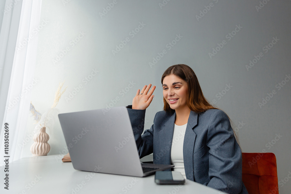 A smiling woman in a gray blazer is looking at her laptop and waving her hand during an online meeting. The workplace is well lit and a vase of dried plants adds aesthetic appeal. Copy space. A smiling woman in a gray blazer is looking at her laptop and waving her hand during an online meeting. The workplace is well lit and a vase of dried plants adds aesthetic appeal. Copy space.