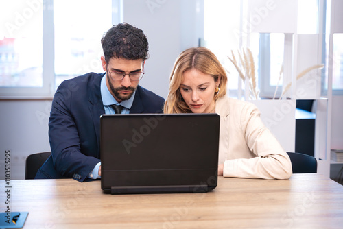 A man and a woman collaborate in a modern office, sharing a laptop screen while working on a project. They smile at each other, showing a productive and friendly business partnership.