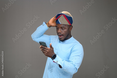 Young Surprised Yoruba Man Looking at His Phone with One Hand on His Head, Dressed in a Native Outfit and Matching Fila Cap photo