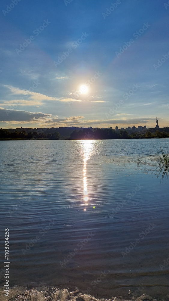 Fototapeta premium Golden sunset reflecting on the calm waters of the Dnipro River, with the Kyiv skyline and Motherland Monument in the distance