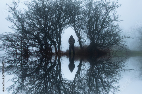 A creepy reflection upside down edit, of a hooded figure, standing in an archway of trees on a dark, eerie winters day in the countryside photo