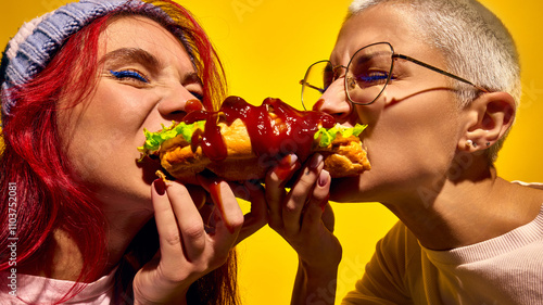 Super delicious street food. Two women emotional and passionately eating hot dog, biting it from two sides against yellow background. Concept of street food, winter fairs, pop art, holiday snacks photo