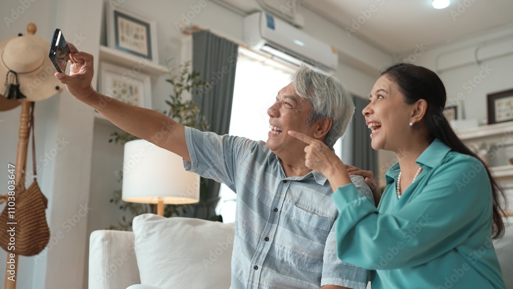 Fototapeta premium Happy grandfather and grandmother making video call with their son at home. Elder man holding smart phone and sitting at sofa while old woman greeting with cheerful vibe at modern house. Myrmidon.