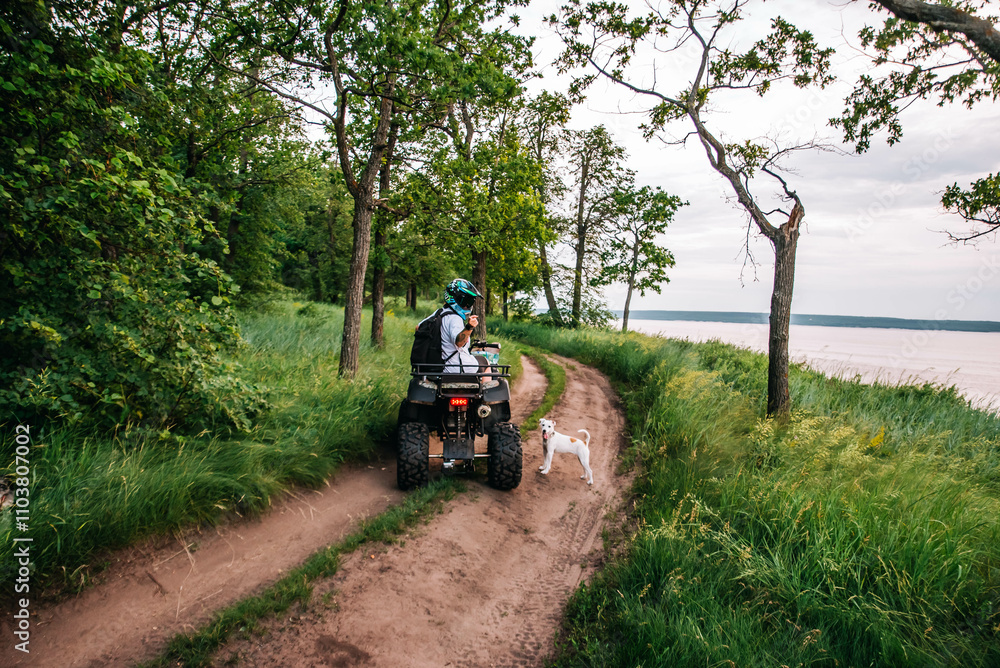 a girl and a guy bikers ride quad bikes in a beautiful green forest on a river cliff, their dogs are running nearby a girl and a guy bikers ride quad bikes in a beautiful green forest on a river cliff, their dogs are running nearby