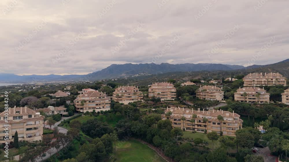 High-angle view of luxurious residential buildings surrounded by greenery and mountains under an overcast sky. High-angle view of luxurious residential buildings surrounded by greenery and mountains under an overcast sky.