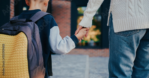 Posterior camera view of little Caucasian boy with blond hair walking towards school together with his mother. Holding hands. Carrying backpack. Starting studying in morning. Education concept.