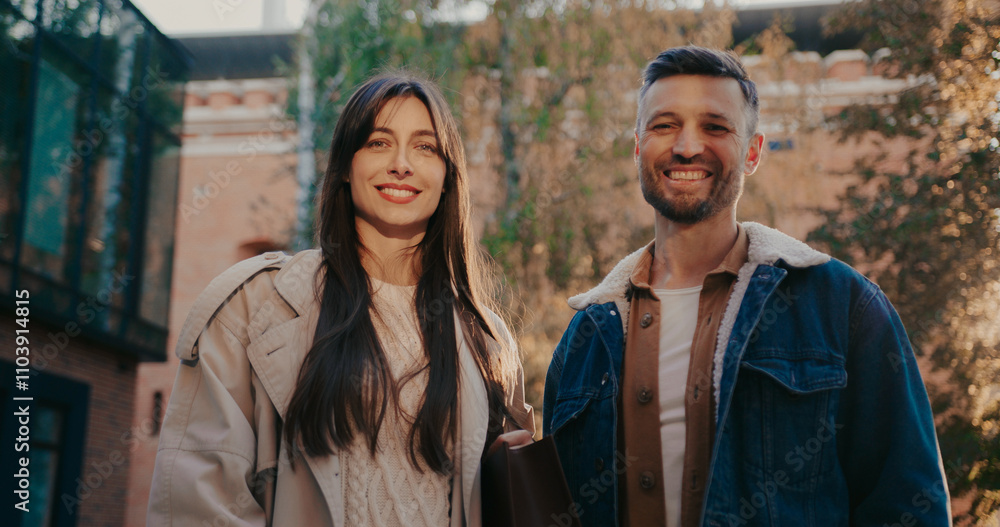 Front view of two people standing together. Looking at each other before glancing at camera. Car parked in front of tall trees in background. Family enjoying fresh air in park. Sunshine.