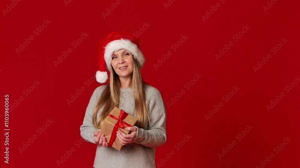 A cheerful woman in a Santa Claus Christmas hat stands on a red background with a gift in her hands. Christmas atmosphere. copy space. A cheerful woman in a Santa Claus Christmas hat stands on a red background with a gift in her hands. Christmas atmosphere. copy space.