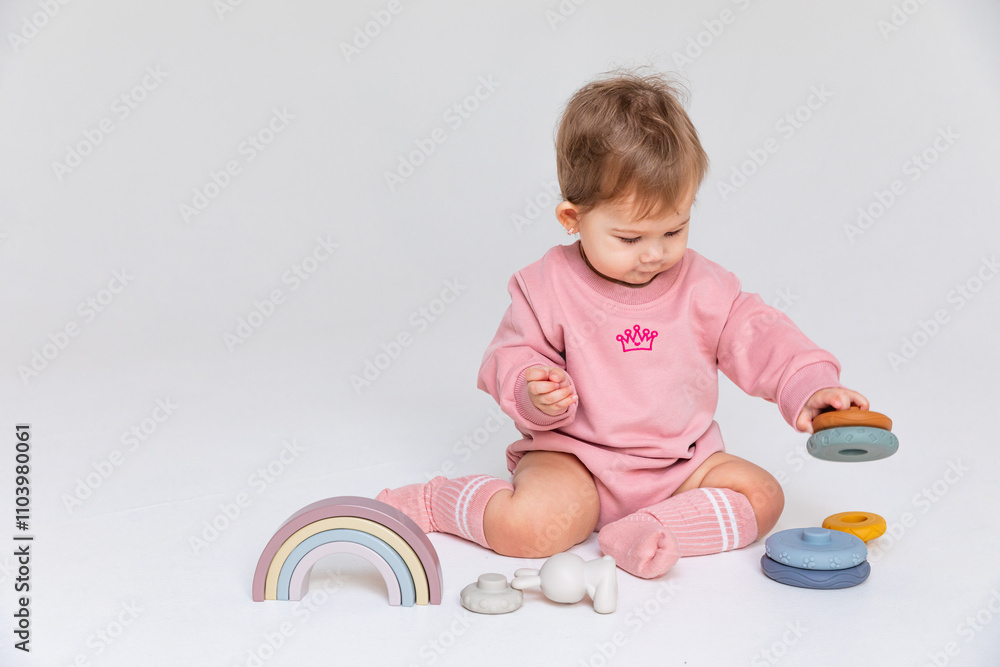 Happy smiling little baby girl sits on the floor and plays with a multi-colored pyramid sorter and rainbow toy on a white background. Development of fine motor skills. Early education. Copyspace Happy smiling little baby girl sits on the floor and plays with a multi-colored pyramid sorter and rainbow toy on a white background. Development of fine motor skills. Early education. Copyspace