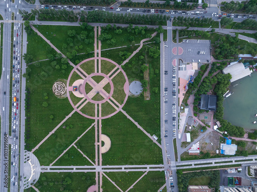 Labyrinth in a children's park, square. Yellow and pink paths for pedestrians among green grass. Bushes and trees on the lawn. Many cars are stuck in a traffic jam on the road.