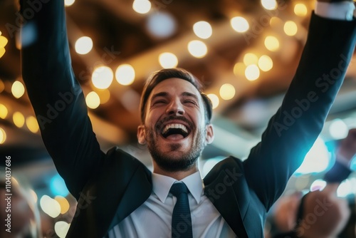 A young man in a suit raises his arms in excitement during a lively celebration at a well decorated venue. Soft ambient lights create a festive atmosphere, showcasing the spirit of joy and camaraderie photo