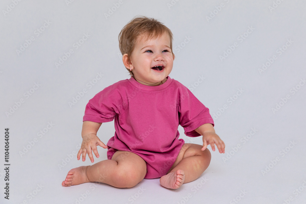 Happy baby girl in a pink bodysuit sitting on a white background and looking at empty copy space. Closeup. Childhood and fashion. Birthday party concept. High quality photo Happy baby girl in a pink bodysuit sitting on a white background and looking at empty copy space. Closeup. Childhood and fashion. Birthday party concept. High quality photo