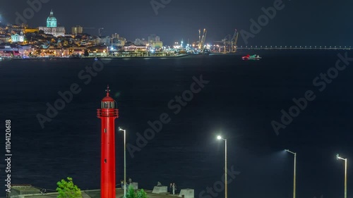 Aerial night timelapse of Cacilhas red lighthouse near bus and railway station with the Tagus River, Lisbon glowing skyline. Boats passing by illuminate the water. Portugal.
