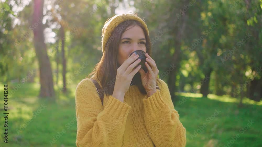 A girl in a yellow sweater and hat drinks tea or coffee from a thermos in a park. Surrounded by green trees and sunset light, she enjoys a peaceful moment outdoors A girl in a yellow sweater and hat drinks tea or coffee from a thermos in a park. Surrounded by green trees and sunset light, she enjoys a peaceful moment outdoors