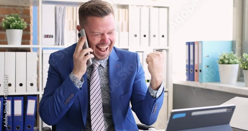 Smiling businessman shakes fist admiringly while talking on modern smartphone. Young entrepreneur receives good news from business partner photo