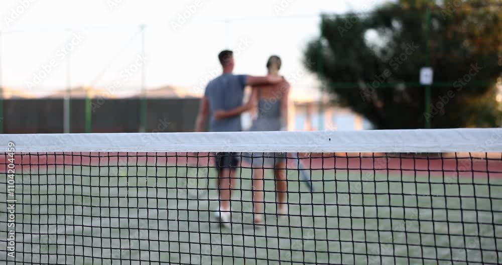 Young man and woman with tennis rackets leave court against net. Young couple hugs reflecting on competitive sports game while cooling down after match Young man and woman with tennis rackets leave court against net. Young couple hugs reflecting on competitive sports game while cooling down after match