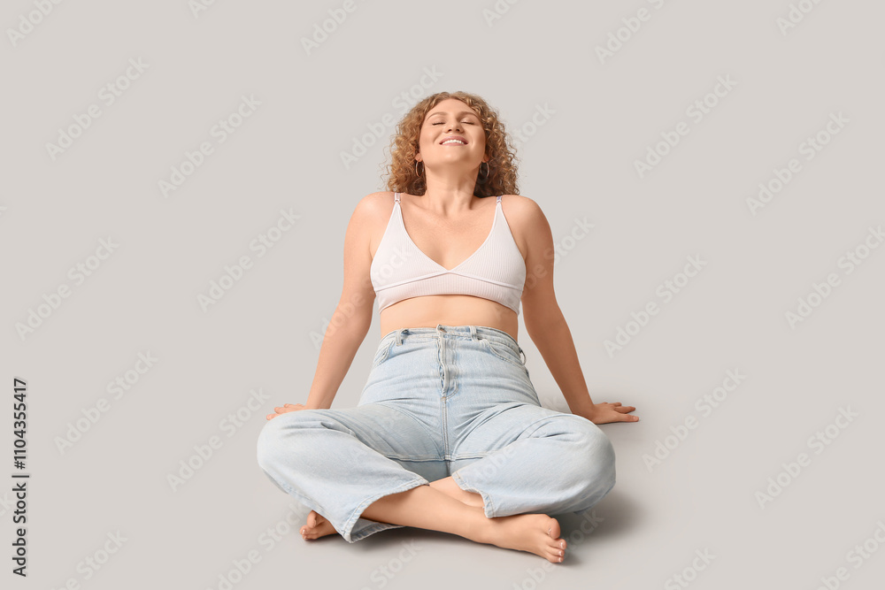 Beautiful young happy body positive woman in stylish jeans sitting on grey background Beautiful young happy body positive woman in stylish jeans sitting on grey background