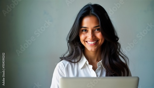 Smiling Indian woman uses laptop. Positive professional portrait. Woman looks happy. Grey background. Business woman on computer. Tech pro. Modern work setting.