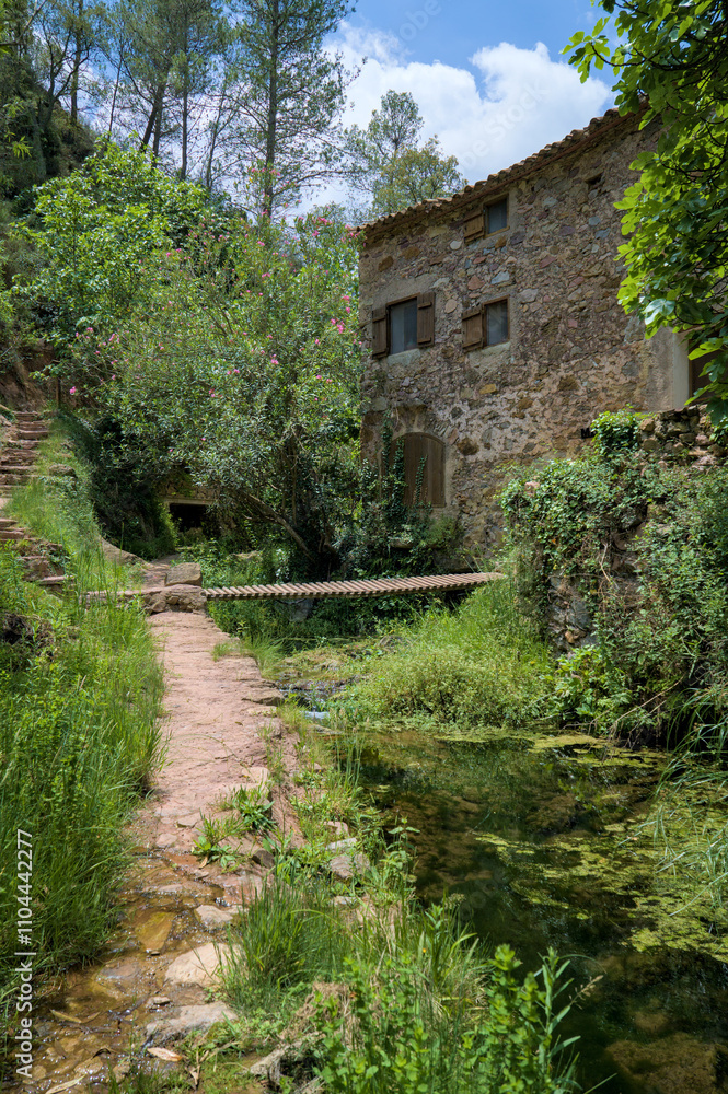 Rustic stone house surrounded by lush greenery, a small wooden bridge crossing a stream. Rustic stone house surrounded by lush greenery, a small wooden bridge crossing a stream.