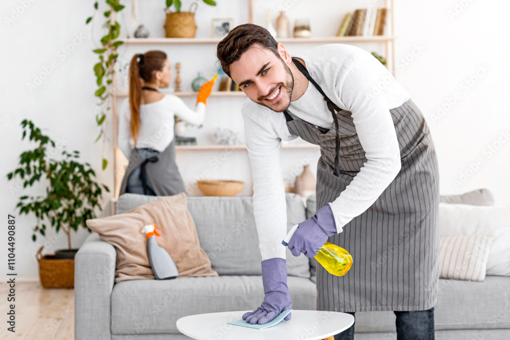 Fototapeta premium Stay at home and cleaning flat spending quarantine together. Cheerful man in apron and rubber gloves with spray and rag wipes dust with wife, near cleaning supplies in interior of living room