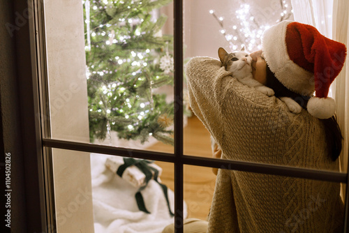 Merry Christmas! Beautiful woman in santa hat in window hugging cute cat on background of festive living room with christmas tree lights. Atmospheric christmas eve at home. Holiday scene