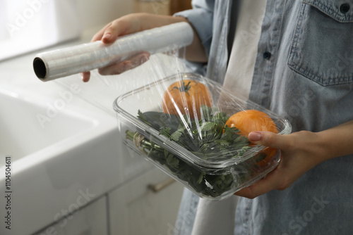 Fotografija Woman putting plastic food wrap over glass container with vegetables in kitchen,