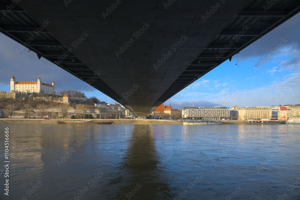 Underneath a large bridge spanning a calm river with a historic castle and cityscape in the background. Underneath a large bridge spanning a calm river with a historic castle and cityscape in the background.