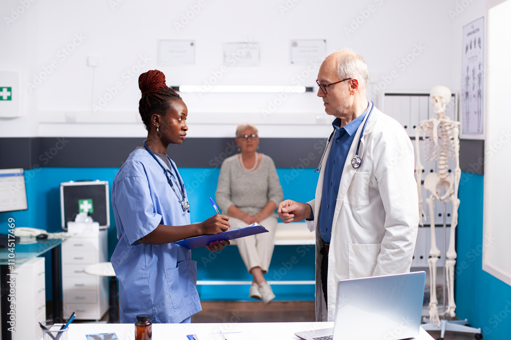 Obraz premium Elderly male physician and female medic assistant are deep in discussion about patient health. Black nurse with clipboard, standing in clinic room with retired doctor and writing medical instructions.