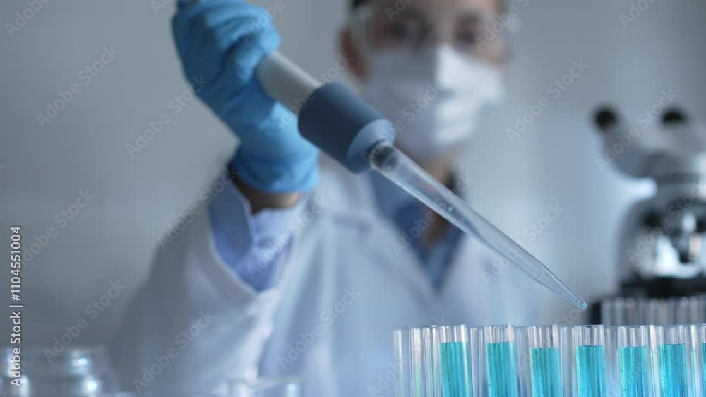 Female scientist researcher wearing a lab coat, mask, and safety glasses is using a micropipette to drop a blue liquid into a test tube, close up view. Medicine and science concept Female scientist researcher wearing a lab coat, mask, and safety glasses is using a micropipette to drop a blue liquid into a test tube, close up view. Medicine and science concept