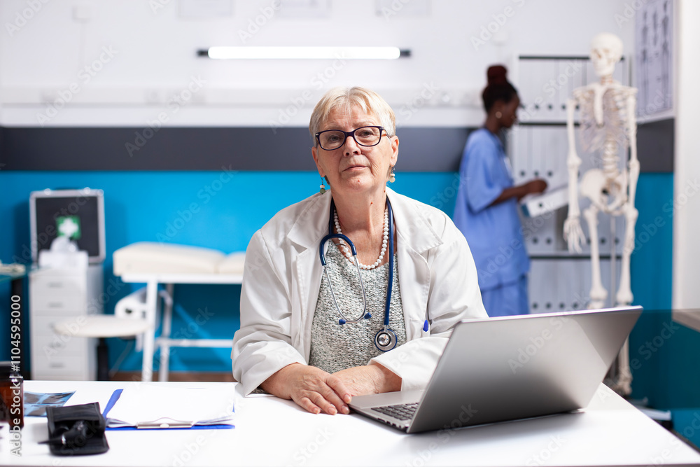 Obraz premium Senior female doctor in lab coat sits at clinic desk with laptop, looking directly at camera. Portrait of elderly physician appearing focused as she prepares for next medical consultation in hospital.