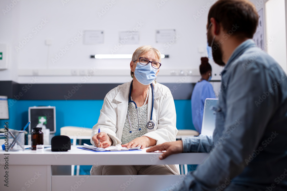 Obraz premium Male patient sharing sickness symptoms with friendly elderly female medic who writes on clipboard during clinic appointment. Senior doctor consults with young man in hospital, both wearing face mask.