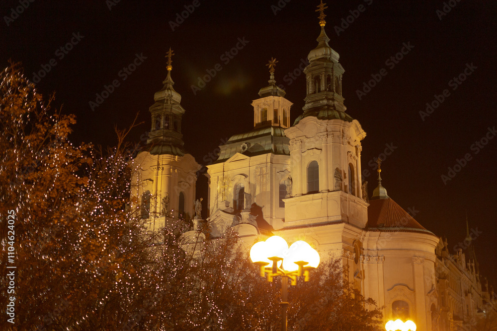 Obraz premium Illuminated historic church towers shine at night, surrounded by vibrant foliage in an urban setting, showcasing intricate architecture and striking lighting that enhance their beauty