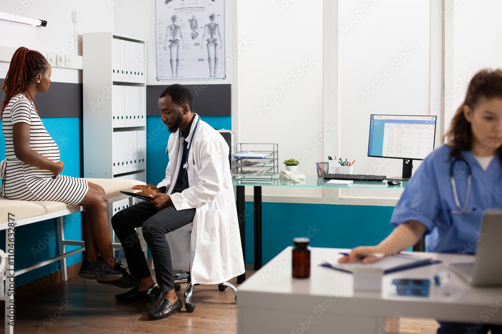 Obraz premium Image showcases multiethnic healthcare professionals using digital devices while pregnant woman sits on hospital bed. African american doctor discusses maternity care plan with young expectant mother.