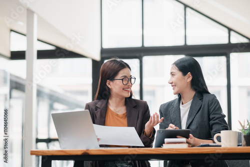 Two businesswomen in a modern office setting, discussing work with documents and a tablet, showcasing teamwork and collaboration.