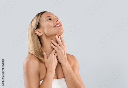 Happy smiling attractive half-naked middle aged blonde woman covered in towel touching her neck, enjoying anti-aging SPA salon treatment results, grey studio background, copy space