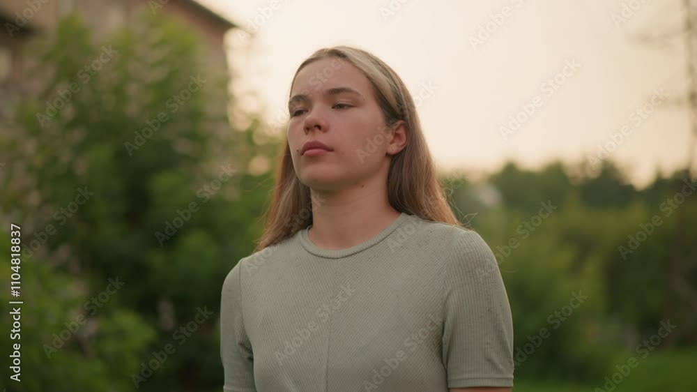 Close-up of young woman walking outdoors with a tired expression, dressed casually, against blurred background of greenery, power lines, and distant building Close-up of young woman walking outdoors with a tired expression, dressed casually, against blurred background of greenery, power lines, and distant building