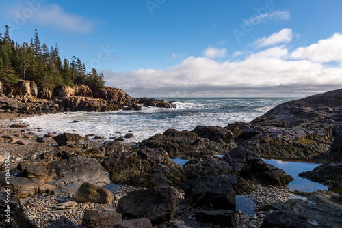 pebble beach in maine blue sky with clouds waves crashing on the rocky east coast beach pines in the background 