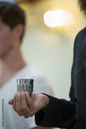 jewish rabbi's hand holding a small silver kiddush cup with wine for blessing of shabat day reciting a traditional Jewish blessing over wine.
